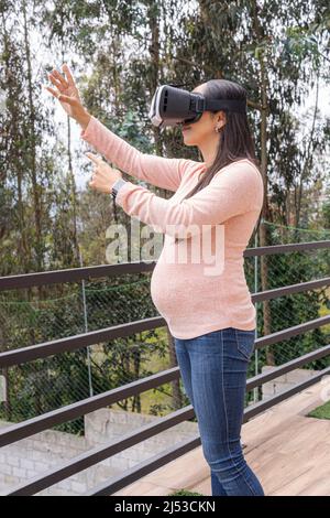 Photo verticale d'une femme latine enceinte sur une terrasse à l'aide de lunettes de réalité virtuelle dans un environnement naturel Banque D'Images