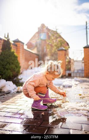 Fille joue dans une flaque avec bâton de bois dans le jour de printemps à la lumière du soleil Banque D'Images