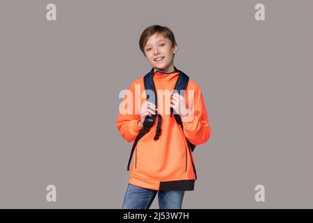 Adolescent de race blanche dans un sweat-shirt orange avec un sac à dos sur son dos posé dans le studio sur un fond gris. Joyeux étudiant souriant nouveau jour Banque D'Images