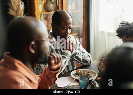 Africain mature homme assis à table avec ses yeux fermés et priant avec sa famille pendant le dîner Banque D'Images