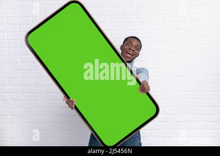 Homme noir excité debout et pointant Big smartphone avec écran vert blanc sur fond de brique blanche, image maquette. Banque D'Images