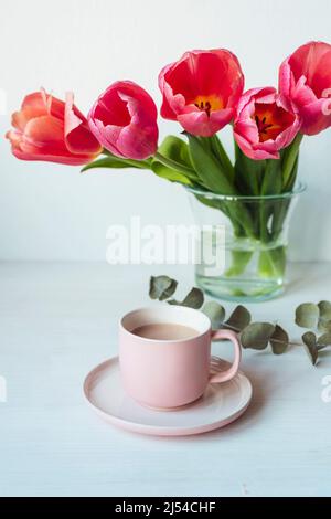 Café dans une tasse rose et bouquet de fleurs de tulipe dans un vase en verre sur une table blanche. Concept vacances de printemps. Gros plan Banque D'Images