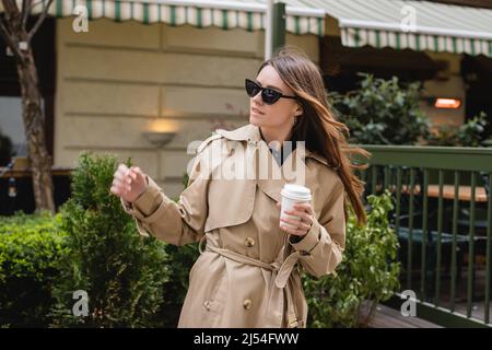 jeune femme en lunettes de soleil tendance et trench coat tenant une tasse en papier Banque D'Images