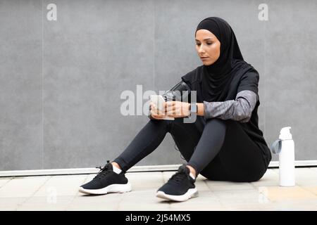 Jeune athlète arabe confiant dans hijab, vêtements de sport avec téléphone et bouteille d'eau, assis sur fond de mur gris Banque D'Images