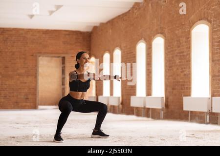Bonne jeune femme arabe brunette athlète avec casque et téléphone, faisant des squats dans le bâtiment de brique Banque D'Images