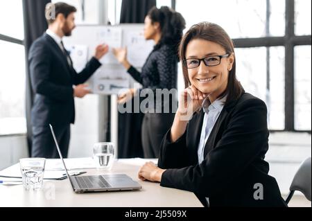 Femme d'affaires caucasienne intelligente avec des lunettes, assise au bureau lors d'une conférence, près d'un ordinateur portable. Belle femme élégante lors d'une réunion d'information, regardant la caméra, souriante et amicale Banque D'Images