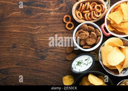 Aliments ou collations malsains. Tous les en-cas classiques de pommes de terre avec des arachides, du pop-corn et des rondelles d'oignon et des bretzels salés dans des assiettes à bol sur fond de bois ancien. Banque D'Images