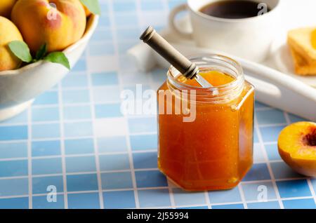 Confiture de pêche maison dans un bocal avec quelques pêches fraîches, une tasse de café et quelques toasts, sur un fond bleu clair. Banque D'Images