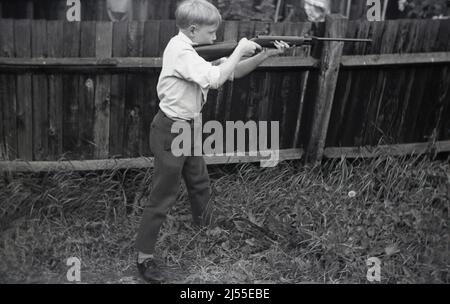 1960s, historique, à l'extérieur dans un jardin à l'arrière, à côté d'une clôture en bois, un garçon debout, pieds à l'écart, tenant une carabine à air comprimé dans les deux mains dans une position de tir ou de tir, peut-être un modèle Diana 27, Angleterre, Royaume-Uni. Banque D'Images