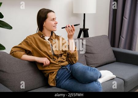 Belle femme attrayante Envoyer un message vocal à l'aide de l'application de messagerie vocale. Inspiré par les innovations. Une jeune femme heureuse assise sur un canapé parle au téléphone et donne Banque D'Images