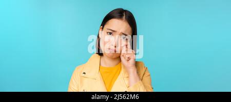 Gros plan de triste fille asiatique pleurant, essuyant des larmes et des tergiverser, regardant bouleversé et sombre, posant sur fond bleu Banque D'Images