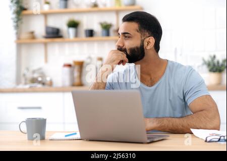 Un homme indien ou arabe attentionné dans un t-shirt décontracté, indépendant, concepteur, travaillant à la maison, assis dans la cuisine avec un ordinateur portable, regardant loin, pensant à un nouveau projet, stratégie Banque D'Images