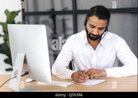 Un homme indien ou arabe intelligent et concentré, un responsable, un entrepreneur, portant un casque, assis dans un bureau moderne, à l'écoute d'un webinaire, d'une conférence, d'améliorer les compétences, de prendre des notes Banque D'Images