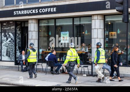 Un trio de travailleurs en vestes haute visibilité passe devant des clients assis à l'extérieur d'une succursale de Starbucks à Margaret Street, Londres, alors que le temps chaud frappe le Royaume-Uni Banque D'Images