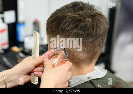 Un adolescent dans un salon de beauté obtient une coupe de cheveux, un coiffeur coupe le garçon d'un adolescent, une coupe de cheveux avec une tondeuse à cheveux et un peigne. Banque D'Images