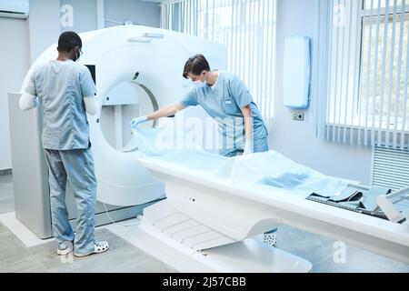 Infirmières multi-ethniques dans les masques et les exfoliants préparation de l'IRM pour l'examen du patient: La femme étalant la feuille jetable sur la table Banque D'Images