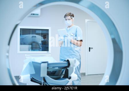 Infirmière sérieuse dans le masque de protection et les gants utilisant la tablette et la lecture de la carte du patient tout en préparant le scanner IRM pour le patient covid Banque D'Images