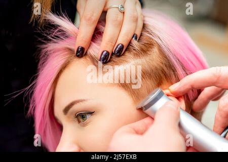 Le coiffeur rasse la femme temple avec des cheveux roses par rasoir électrique dans un salon de coiffure, gros plan Banque D'Images