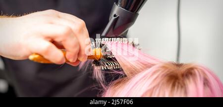 Séchage de cheveux courts roses de jeune femme caucasienne avec un sèche-cheveux noir et une brosse ronde noire par les mains d'un coiffeur de sexe masculin dans un salon de coiffure, gros plan Banque D'Images