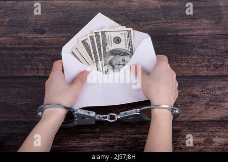 Les mains menottées tiennent une enveloppe blanche ouverte avec des dollars sur un fond en bois sombre Banque D'Images