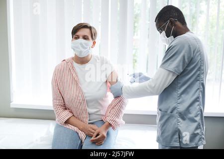 Infirmière afro-américaine concentrée dans le masque donnant le vaccin Covid au patient sur le canapé à l'hôpital Banque D'Images