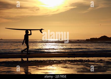 garçon marchant au coucher du soleil sur la plage solitaire portant sa planche de surf Banque D'Images