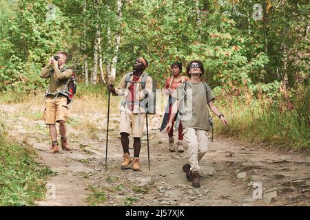Groupe de jeunes amis interraciaux avec des sacs à dos en randonnée tout en marchant le long du chemin dans la forêt et en regardant autour Banque D'Images