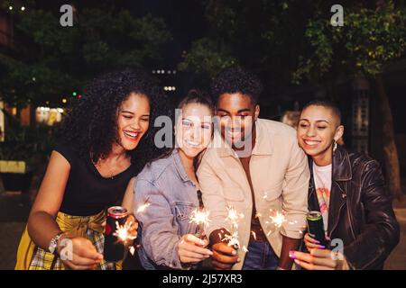 Amis célébrant la saison des fêtes avec des lumières bengale. Groupe de jeunes heureux tenant des canettes de sparkers et de bière la nuit. Amis joyeux de la ville Banque D'Images