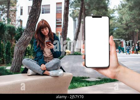 La main d'une femme tient un smartphone avec un écran blanc, gros plan. Une jeune femme est assise sur un banc et utilise un smartphone. Le concept de communication en ligne Banque D'Images