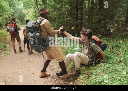 Randonneur afro-américain avec sac à dos donnant la main à l'homme tombé tout en l'aidant à se lever pendant la randonnée en forêt Banque D'Images