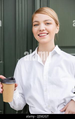 femme blonde heureuse avec un verre à emporter souriant à l'extérieur Banque D'Images