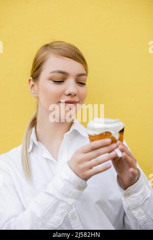 femme blonde en chemise blanche tenant un cupcake flou isolé sur jaune Banque D'Images