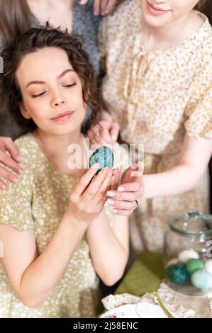 Trois sœurs belles jeunes femmes prêtes à célébrer les vacances de printemps de joyeuses Pâques à la maison décorant table à manger avec des oeufs de Pâques tenant Banque D'Images