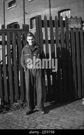 1950s, historique, un jeune homme en travail combinaisons debout à l'extérieur de la porte en bois de locaux industriels et de son lieu de travail, Angleterre, Royaume-Uni. Banque D'Images