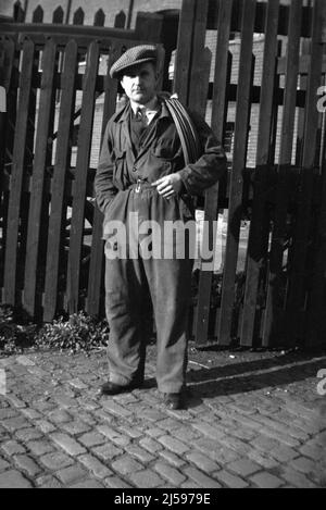1950s, historique, un homme en travail combinaisons et coth cap, tenant un rouleau de câblage, debout à l'extérieur de la porte en bois vers les locaux industriels et son lieu de travail, Angleterre, Royaume-Uni. Banque D'Images
