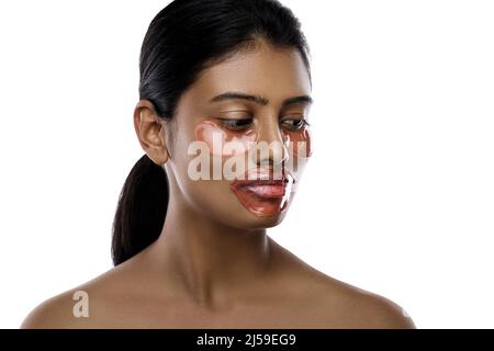 Belle femme indienne avec des timbres hydratants pour les yeux et un masque de lèvre sur son visage sur fond blanc. Banque D'Images