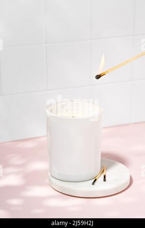Une bougie aromatique faite de cire de soja est illuminée avec un match. Bougie écologique et végétalienne sur une table rose. Aromathérapie, parfum, détente. Banque D'Images