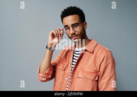 Portrait d'un jeune homme noir à la mode plein de confiance en chemise corail qui règle les lunettes sur fond gris Banque D'Images
