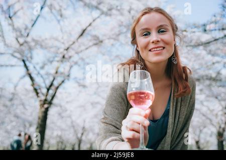 Portrait d'une femme souriante profitant de la nature au printemps. Pique-nique sous les cerisiers en fleurs. Femme profitant de la vie, boire du vin, offrant un verre de vin à Banque D'Images