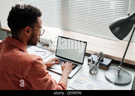 Vue sur l'épaule d'un jeune gestionnaire noir ciblé dans des vases assis à un bureau devant des stores fermés et à la recherche d'informations sur un ordinateur portable Banque D'Images