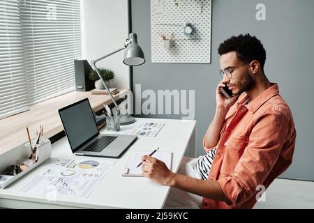 Jeune homme de course mixte sérieux avec barbe de 3 jours assis au bureau avec un ordinateur portable et des papiers et planification de la réunion tout en parlant au client par téléphone Banque D'Images