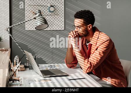 Un jeune homme noir perplexe dans une chemise de corail assis avec les mains classed à la table et regardant l'écran d'un ordinateur portable tout en pensant à un problème d'affaires Banque D'Images