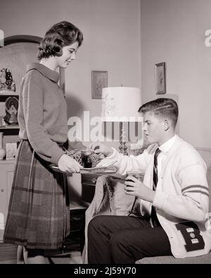 1960S DATANT JEUNE COUPLE DANS LA SALLE DE SÉJOUR FILLE SERVANT PLATEAU AVEC COLA ET BRETZELS À SON PETIT AMI PORTANT UNE VARSITY CHANDAIL - J1484 HAR001 HARS BIENVENUE COLA STYLE DE VIE FEMMES SANTÉ MAISON VIE COPIE ESPACE AMITIÉ DEMI-LONGUEUR PERSONNES PETIT AMI HOMMES ADOLESCENT GARÇON B&W RENCONTRES PETITE AMIE BUTS ENCAS BONHEUR LOISIRS ET FIERTÉ ATTRACTION CRAVATE CHANDAILS VARSITY CONNEXION COUR PRETZELS LETTRE CHANDAIL ÉLÉGANT POSSIBILITÉ DE CROISSANCE ADOLESCENT ACTIVITÉ SOCIALE TOGETHERNESS NOIR ET BLANC CAUCASIEN ORIGINE ETHNIQUE COURTING HAR001 DÉMODÉE Banque D'Images