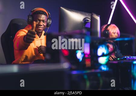 Joyeux Afro-américain dans un sweat à capuche orange assis dans un cyber café, gagnant un autre match avec ses compagnons, montrant les pouces jusqu'à la caméra. Photo de haute qualité Banque D'Images