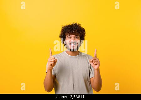 Homme indien attrayant avec une expression faciale heureuse pointant vers le haut avec les deux doigts, présentant l'espace de copie. Studio d'intérieur isolé sur fond jaune Banque D'Images
