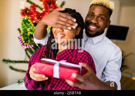 un bel homme brésilien garde les yeux de sa petite amie couverts pendant qu'il donne un cadeau tenant boîte cadeau célébration noël à la maison Banque D'Images