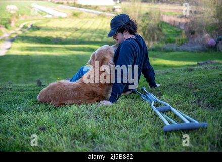 Des béquilles au premier plan de cette ferme rurale idyllique scène avec un jeune homme et son chien d'or affectueux qui se câblent avec un aroun de bras Banque D'Images