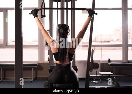 Machine à abaisser l'épaule. Une femme de fitness s'entraîne dans la salle de gym. Exercice de force du haut du corps pour le haut du dos. Vue du dos sans visage Banque D'Images