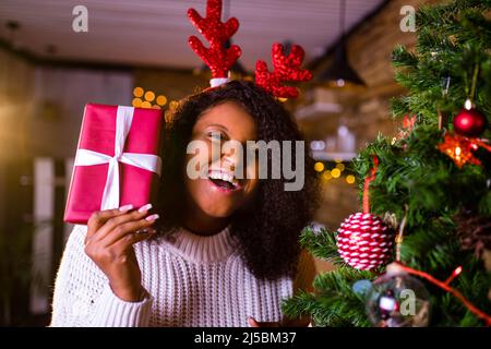 brésil brunette cheveux femme tenir la boîte cadeau rouge près de sapin-arbre à son appartement elle porte le chandail blanc et les cornes de cerf rouge Banque D'Images