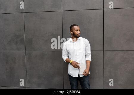 Un jeune homme d'affaires sérieux et confiant est debout à l'extérieur dans un maillot blanc, regardant sur le côté. Un journaliste indépendant afro-américain plein de style Banque D'Images